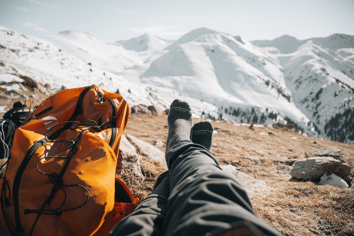 A hiker resting in merino wool socks beside an orange pack on a snowy ridge